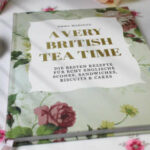 A very british Teat Time liegt auf einer mit Rosen bestickten Tischdecke. Daneben liegt eine rosafarbene Rose sowie zwei silberne Löffel und ein silbernes Teesieb.