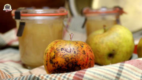 Ein fleckiger Apfel liegt auf einem karieten Geschirrtuch. Im Hintergrund stehen Gläser mit Apfelmarmelade