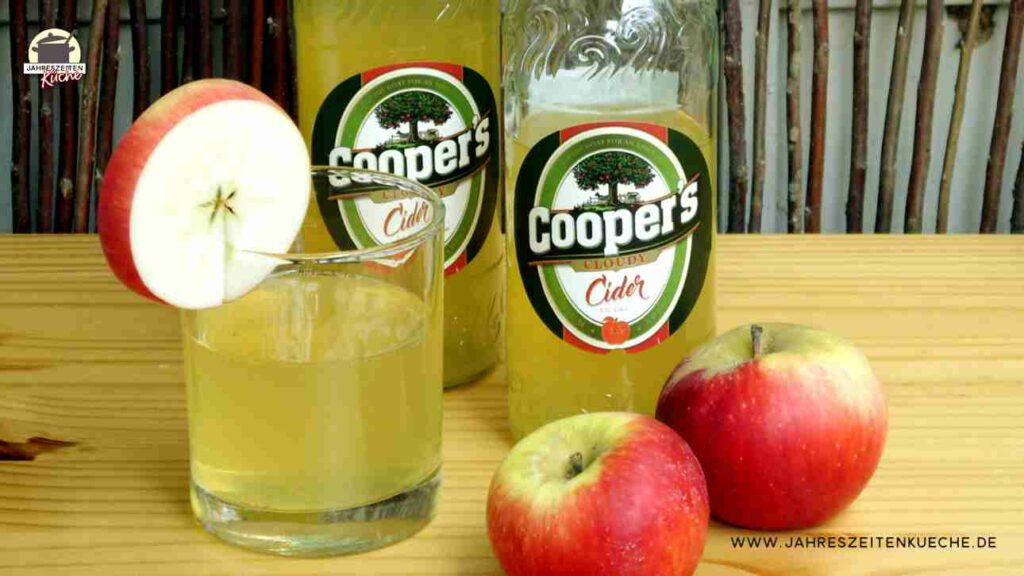 Zwei Flaschen Coopers Cloudy Cider mit einem Glas Cider und zwei Äpfeln auf einer Holzplatte