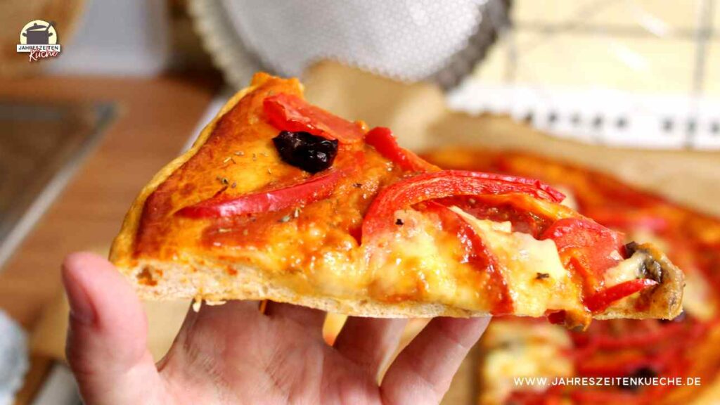 Eine Hand hält ein Stück Dinkelpizza mit Paprika, Oliven und Mozzarella.