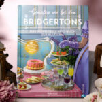 Das Kochbuch Genießen wie bei den Bridgertons ist eingerahmt von einem alten Kerzenleuchter und einem Stapel Bücher, auf denen weiße Handschuhe und eine rote Rose liegen.