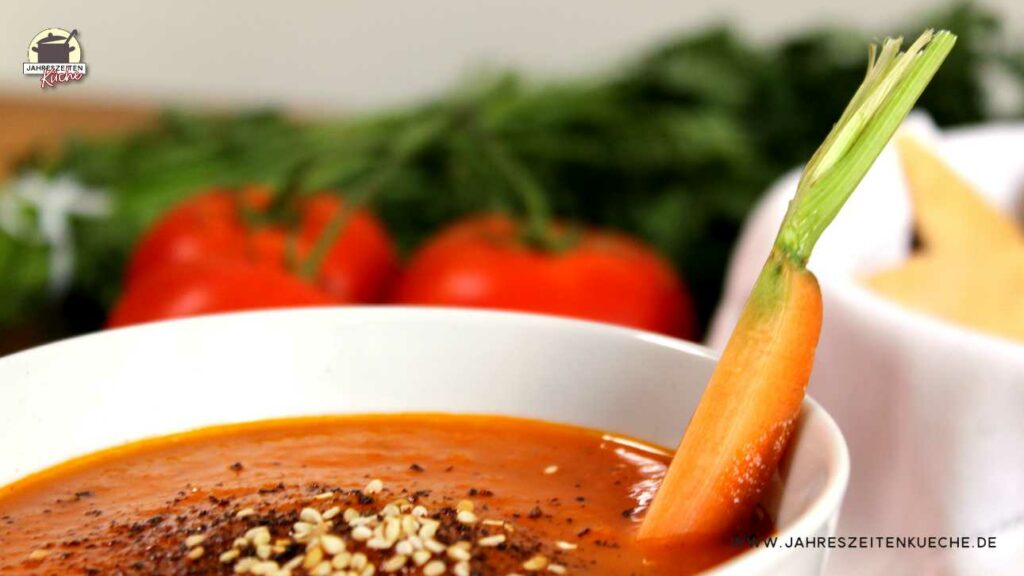 Karotten-Tomaten-Suppe mit Sesam bestreut in einer weißen Porzellanschale. In der Suppe steckt eine Karotte und im Hintergrund frische Tomaten.