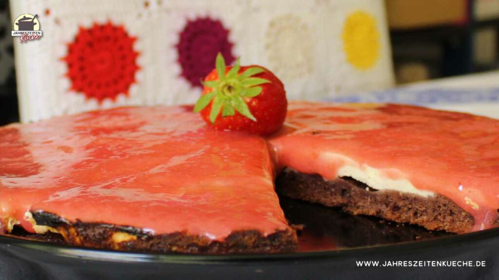 Marzipan-Käsekuchen mit Erdbeermark auf einer schwarzen Kuchenplatte mit einer Erdbeere gekrönt. Ein Stück fehlt.