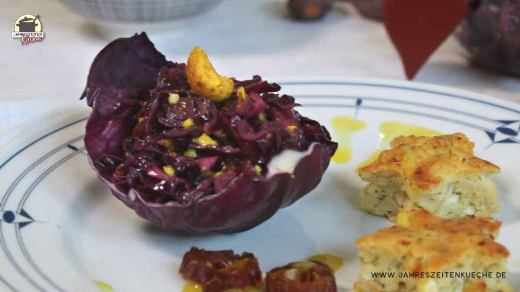 Auf einem Teller liegt ein Rotkohlblatt, in dem Rotkohlsalat mit Cashewkernen angerichtet ist. Daneben liegen Hüttenkäsepuffer.