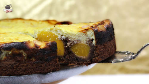 Auf einem Tortenheber liegt ein Stück Schokokuchen mit Pfirsich und Käsekuchendecke.