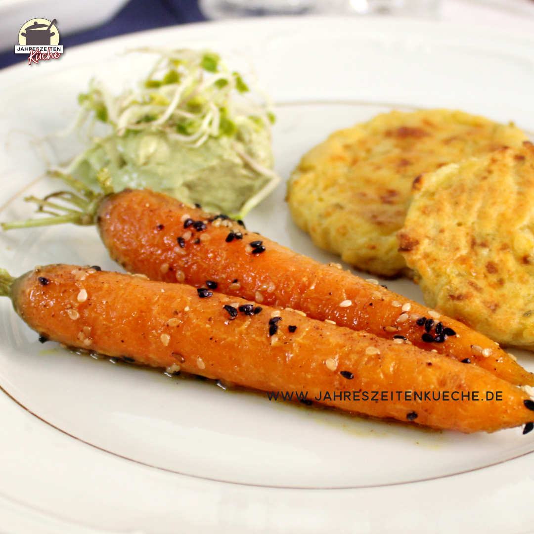 Mit Honig glasierte Karotten liegen auf einem weißen Teller. Im Hintergrund sind Hüttenkäsepuffer und ein veganer Dip