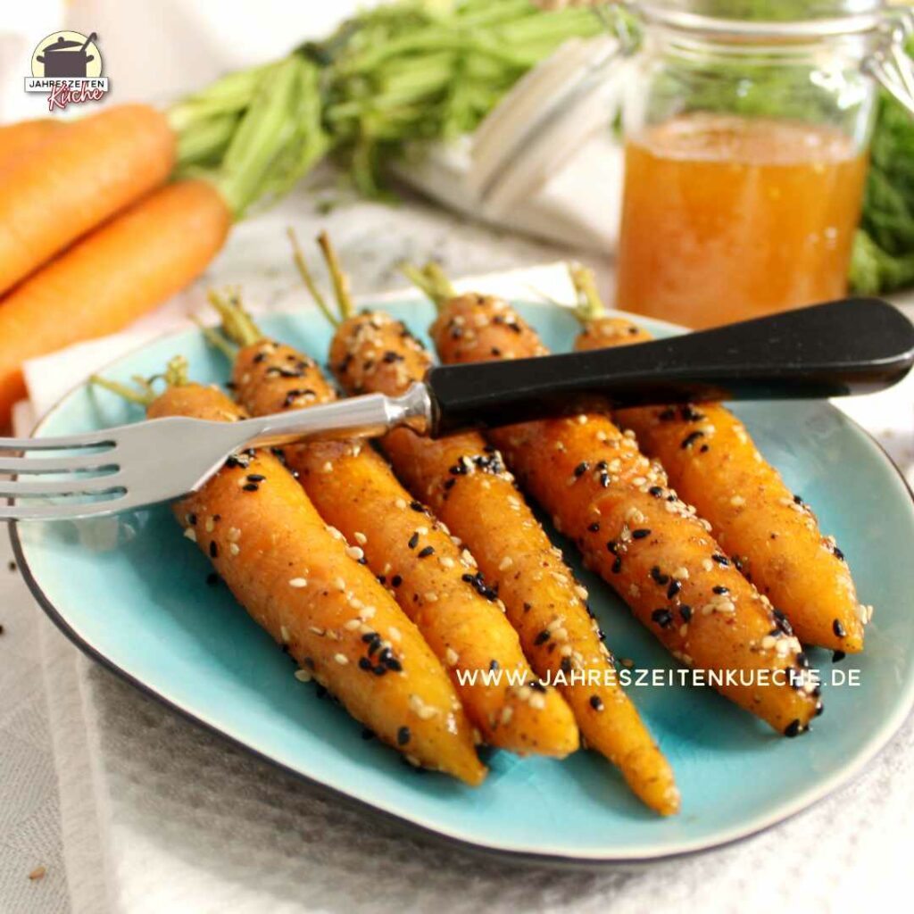 Auf einem türkisfarbenen Teller liegen 5 glasierte Karotten. Quer darüber liegt eine Gabel und im Hintergrund sind frische Möhren