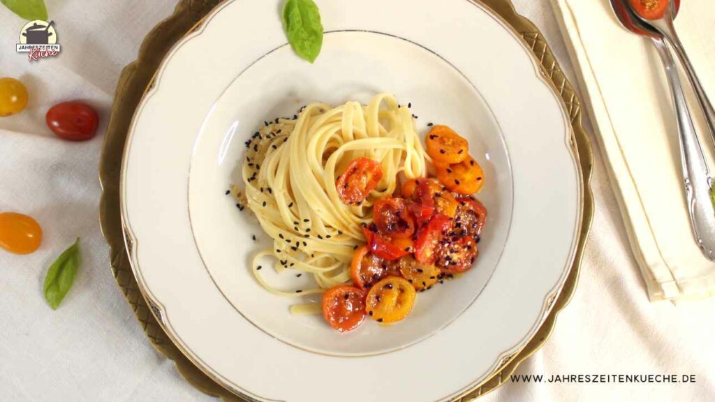 Ein weißer Teller, der auf einem weißene Tischtuch steht, sind Fettuccine und glasierten Honig-Tomaten angerichtet.