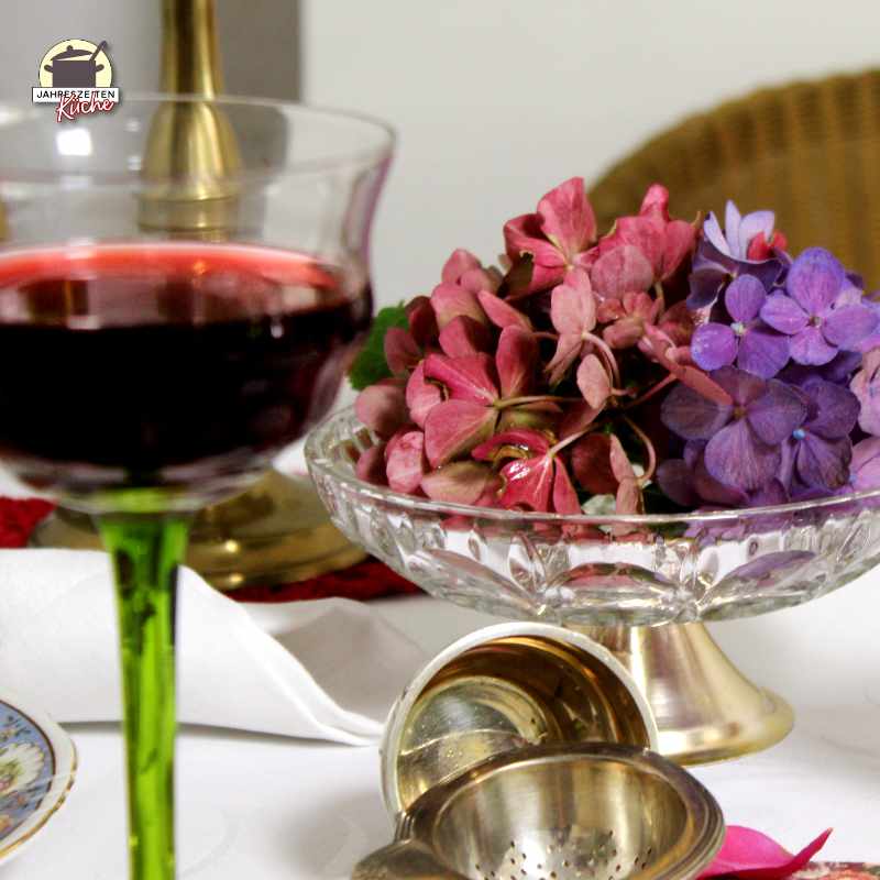 Eine Schale Hortensien, vor der ein silbernes Teesieb liegt und ein Rotweinglas steht.