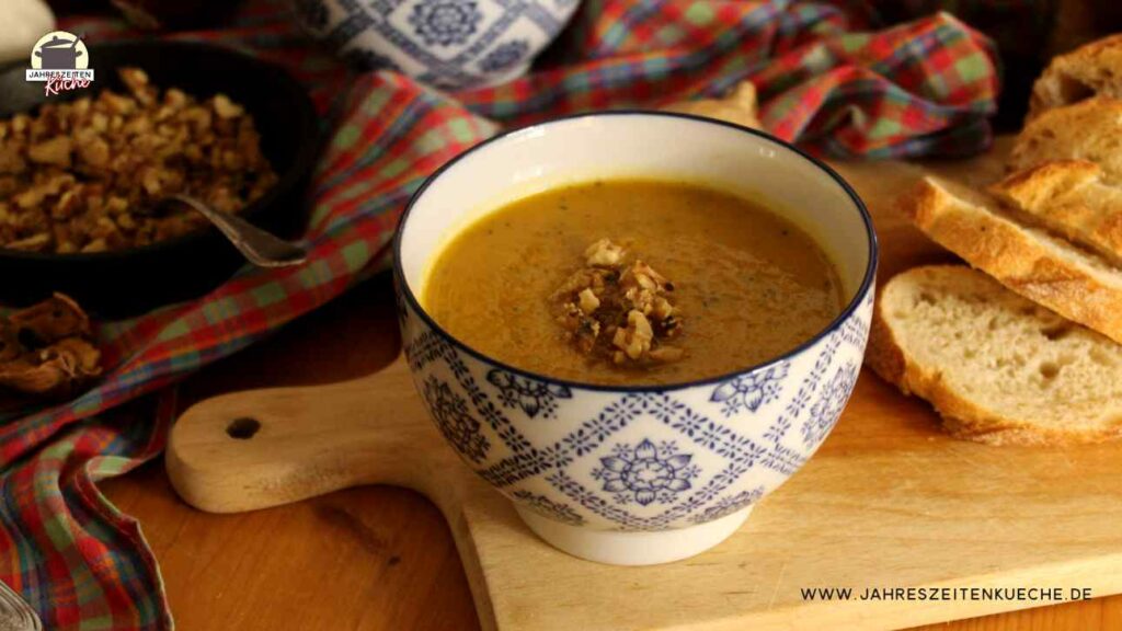 Ein Schälchen mit Karotten-Zucchini-Suppe steht auf einem Holztisch. Im Hintergrund ist ein Pfännchen mit Walnuss-Crunch.