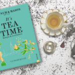 Das Kochbuch It's Tea Time von Tom Parker Bowles liegt auf ausgestreutem Tee. Rechts daneben stehen eine Tasse mit Tee, ein silbernes Teesieb und eines silbernes Kännchen