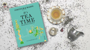Das Kochbuch It's Tea Time von Tom Parker Bowles liegt auf ausgestreutem Tee. Rechts daneben stehen eine Tasse mit Tee, ein silbernes Teesieb und eines silbernes Kännchen