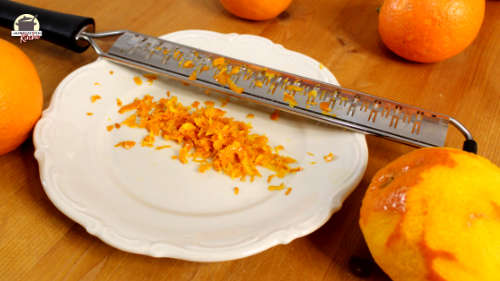 Auf einem Holztisch steht ein Teller mit abgeriebener Orangenschale und einer Reibe. Dahinter liegen orangen.