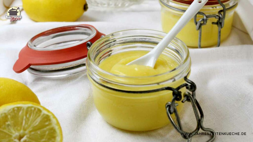 In einem Glas Lemon Curd steckt ein weißer Porzellanlöffel. Im Hintergrund ist ein weiteres Glas und eine Zitrone zu sehen.