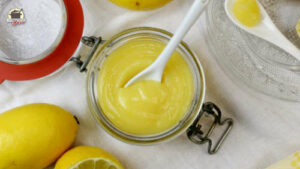 In einem Glas Lemon Curd steckt ein weißer Porzellanlöffel. Daneben liegen Zitronen.
