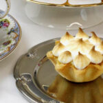 Auf einem silbernen Teller ist ein Lemon-Meringue-Pie. Links daneben steht eine Tasse Tee.