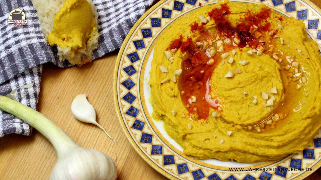 Ein Teller mit Hummus mit Erdnüssen steht auf einem Holztisch. Daneben liegt Knoblauch