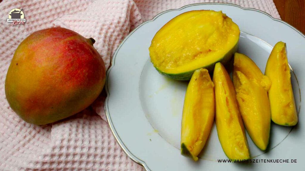 Eine aufgeschnittene Mango liegt auf einem weißen Teller. Daneben liegt eine rötliche Mango