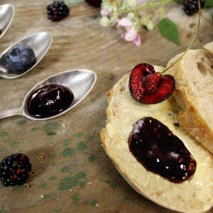 Neben silbernen Teelöffeln, auf denen ein Klacks Brombeer-Heidelbeer-Kirsch-Marmelade mit Rosenblüten ist, liegt eine Scheibe Weißbrot mit einer halbierten Kirsche.