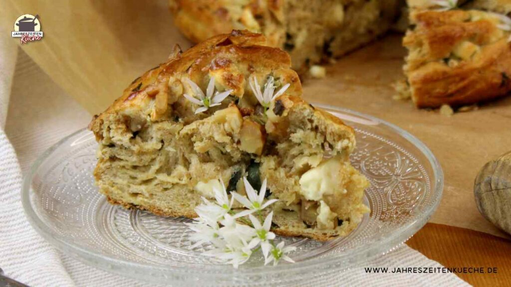 Ein Stück Bärlauchkranz mit Feta und Walnüssen ist mit Bärlauchblüten garniert und liegt auf einem Glasteller