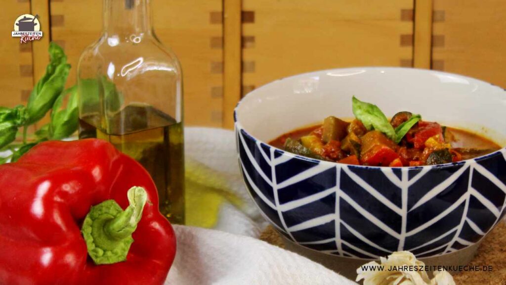 Eine Porzellanschale mit französicher Ratatouille. Im Vordergrund liegt eine rote Paprikaschote