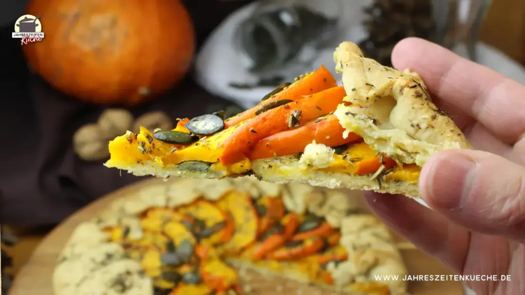 Eine Hand hält Ein Stück Kürbis-Galette. Im Hintergrund ist ein holzteller mit der Kürbis-Galette zu sehen.