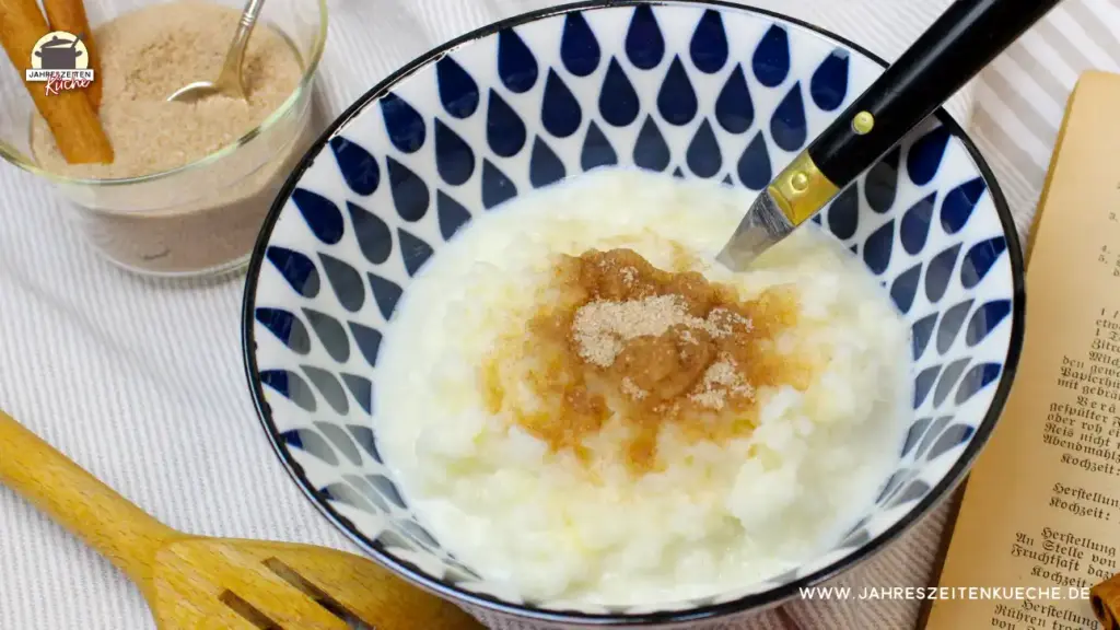 Eine blau-weiß gemusterte Schale in der Milchreis mit Zimt und Zucker ist. Dahinter steht eine Glasschale mit einer Zimt-Zucker-Mischung.