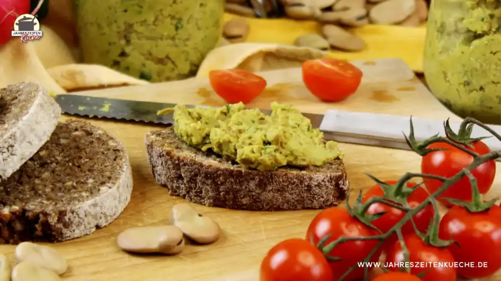 Eine Scheibe Schwarzbrot, die mit Ackerbohnenaufstrich bestrichen ist. Davor liegen kleine Tomaten