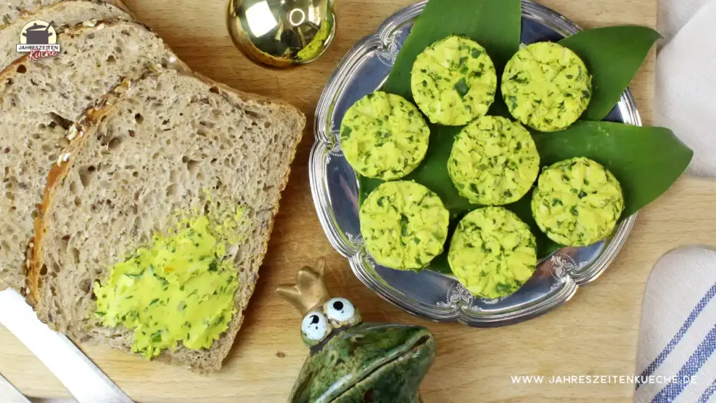 Neben Brotscheiben liegt auf einem silbernen Teller Bärlauchbutter. Darunter eine Frosch-Figur und darüber eine goldene Kugel