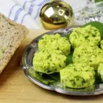 Neben Brotscheiben liegt auf einem silbernen Teller Bärlauchbutter. Dahinter ist eine goldene Kugel