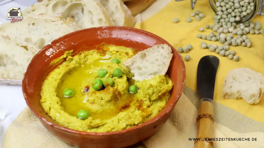 Erbsenhummus in einer Schale mit Olivenöl und Weißbrot angerichtet. Im Hintergrund liegen getrocknete Erbsen