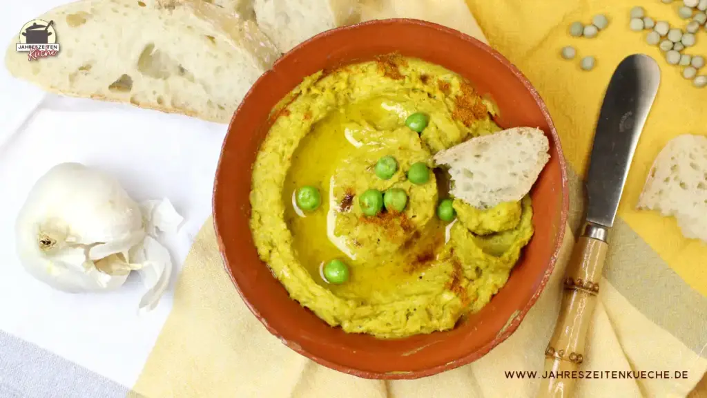 Eine Schale mit Erbsenhummus. Daneben liegt Koblauch, frisches Ciabatta und ein Messer.